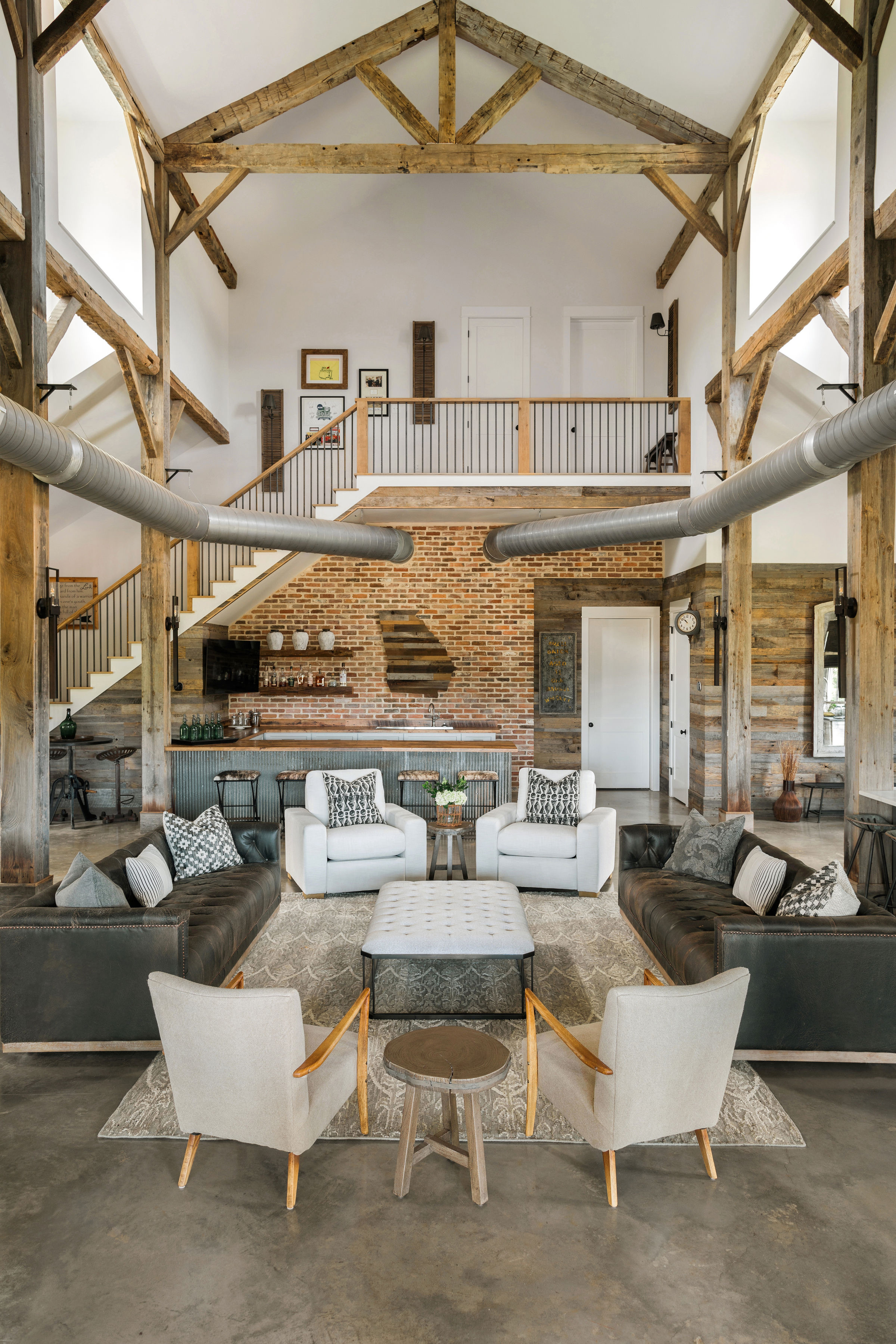Cathedral barn great room with reclaimed timber trusses, brick wall, leather sofas, and mezzanine